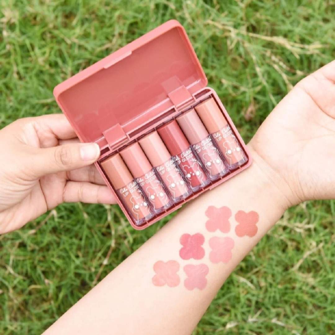 Set of lip glosses in a pink case held by a person with swatches on their wrist against a grass background